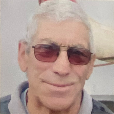 Older man with grey hair, wearing sunglasses and a grey polo shirt, smiling.