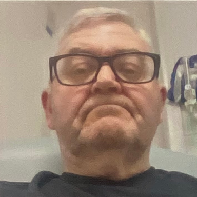 Older man with glasses, frowning and sitting in a clinical setting.