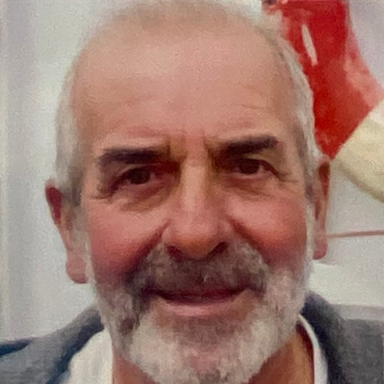 Smiling elderly man with a grey beard, wearing a grey sweater.