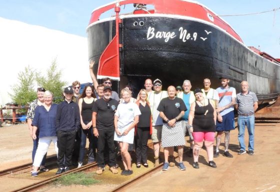 Barge no 9, out of the water, painted black and red, with a large group of smiling volunteers in front of her.